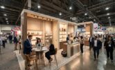 An elevated, photorealistic view of a sprawling ASD Market Week trade show floor at the Las Vegas Convention Center in 2026, where a central custom-designed booth made of warm wood and beige panels serves as a bustling hub. Two professionals, seated at a modern round table with documents, are deep in discussion, while others staff a clean counter with product displays, engaging with diverse attendees. The wide aisle is crowded with visitors from varying backgrounds carrying bags and moving among other distant exhibits under a complex ceiling with visible industrial rigging and ample overhead lighting, all set within a sophisticated, open business environment.