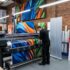 A professional print shop production floor showing a technician operating a large-format printer that is outputting a vibrant, colorful trade show graphic banner. Rolls of printing substrates and rigid panels line the brick wall in the background.