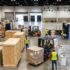 A high-angle, realistic view of a bustling convention center hall during trade show setup. Logistics workers wearing high-visibility yellow vests operate forklifts and assemble booth structures. Large wooden shipping crates, shrink-wrapped pallets with booth number labels, and open loading dock doors with freight trucks are visible in the background, illustrating the complex process of material handling and drayage.
